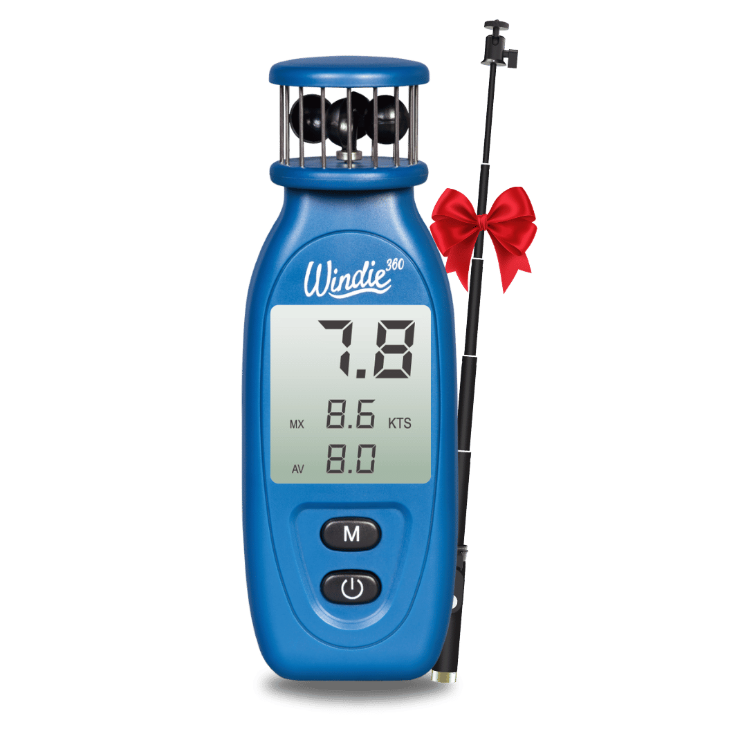 windie.pro blue handheld anemometer displaying wind speed, held by a person on a boat with sailboats in the background.