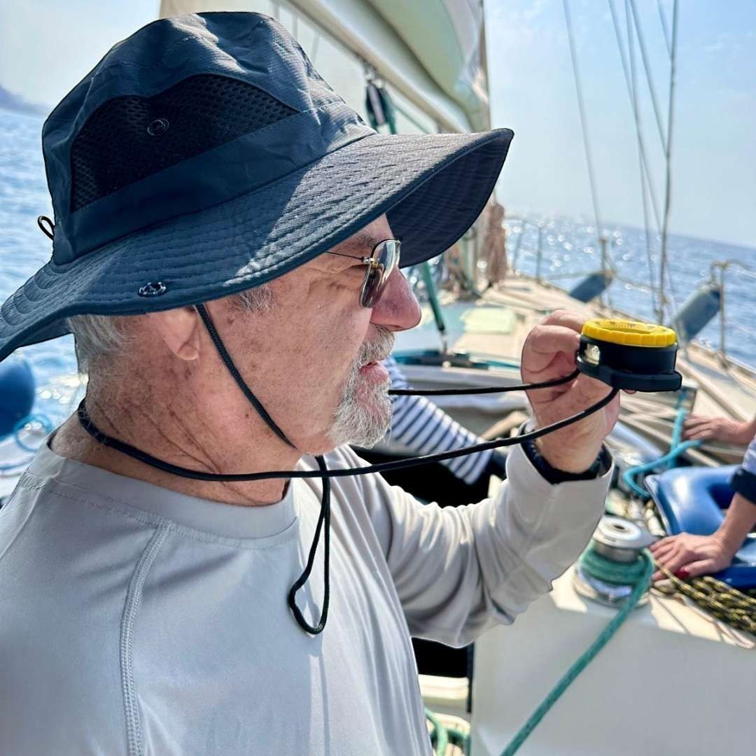 Man on sailboat wearing a dark blue hat and grey shirt, holding a yellow and black wind speed indicator. windie.pro