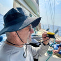 Man on a sailboat holding a yellow and black anemometer and compass set. He wears a dark grey hat and sunglasses.