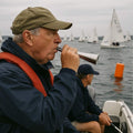 Man in navy jacket and orange life vest blows a silver fog horn on a boat, with sailboats in the background.