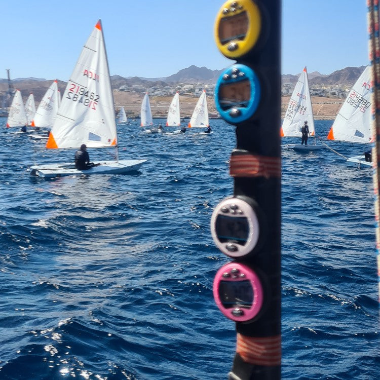 Windie.pro 4-unit timer pack featuring yellow, blue, white, and pink sailing stopwatches attached to a mast, with sailboats in the background.