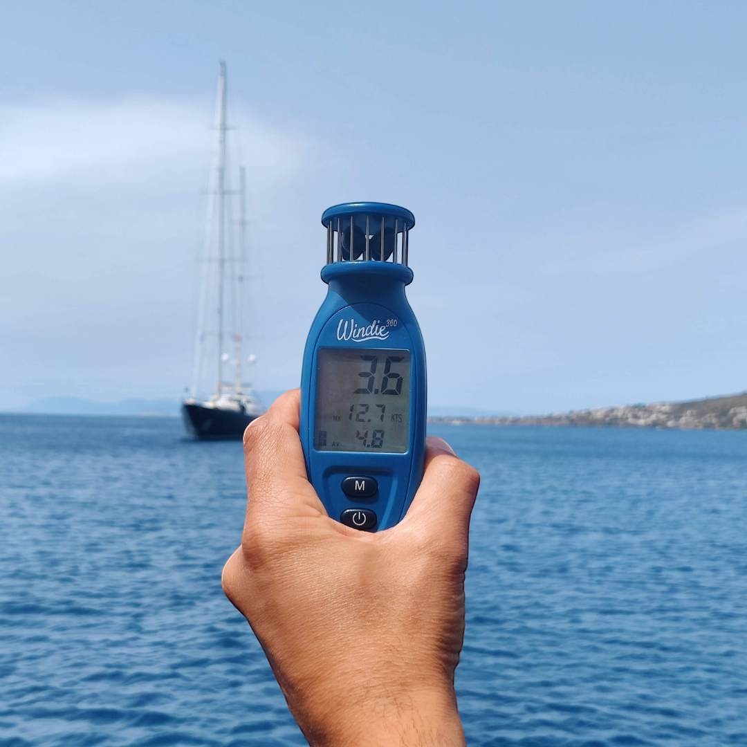 Windie.pro blue handheld digital wind meter displaying wind speed, held in a hand with a sailboat in the background on the water.