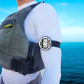 windie.pro sailing timer with digital display on arm, worn by person in life vest over white long-sleeve shirt, blue sky and ocean background.