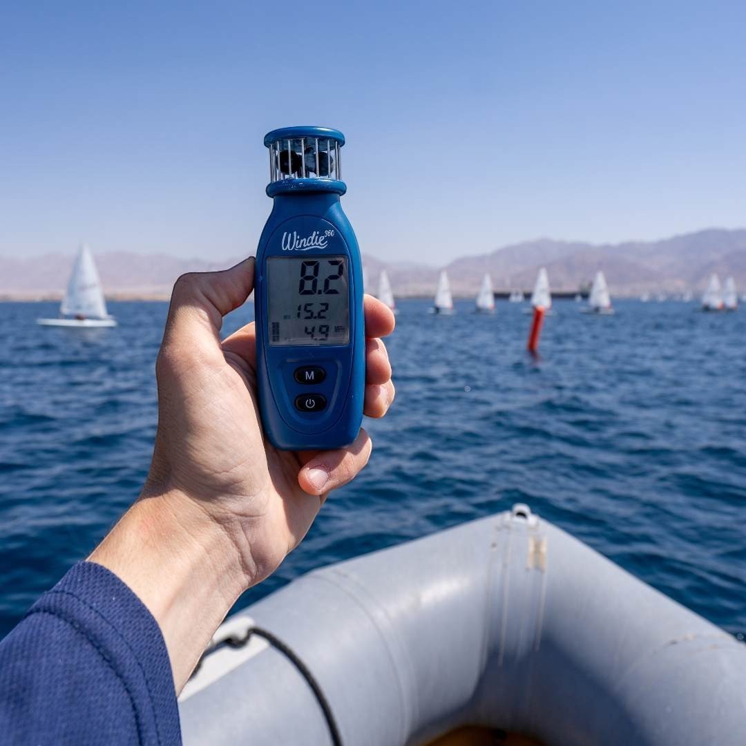windie.pro blue handheld anemometer displaying wind speed, held by a person on a boat with sailboats in the background.