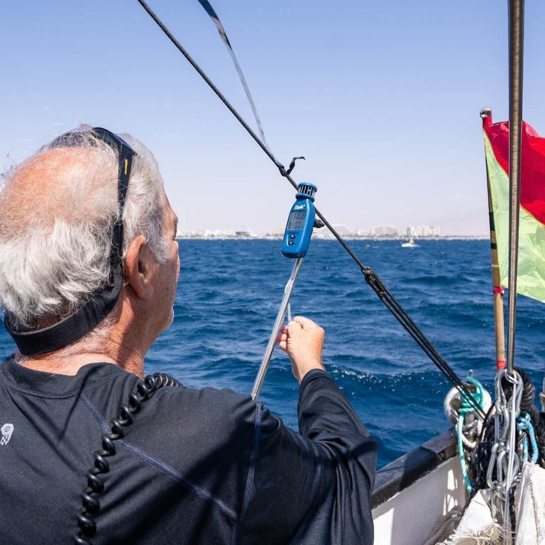 Windie.pro blue handheld wind meter with digital display attached to a sailboat rigging, used by a man in black shirt.